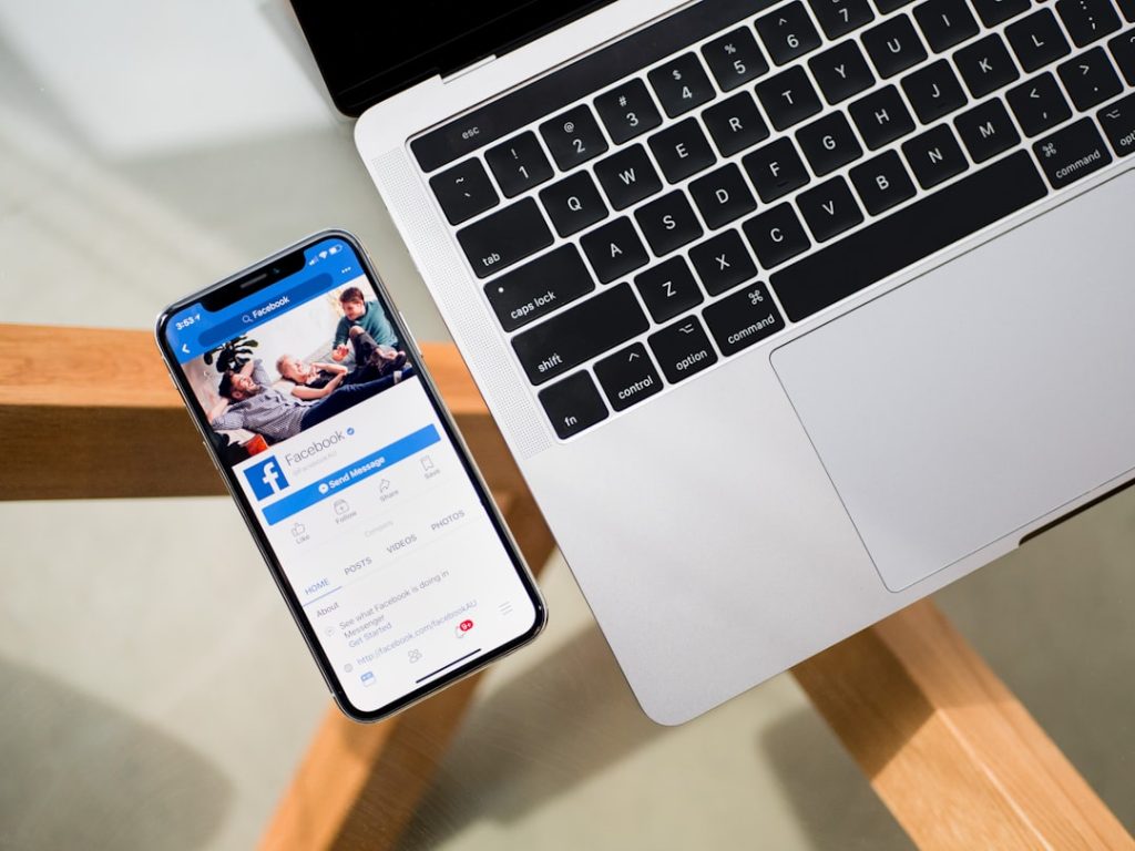 Smartphone displaying the Facebook app open beside a silver laptop on a glass table.