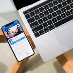 Smartphone displaying the Facebook app open beside a silver laptop on a glass table.