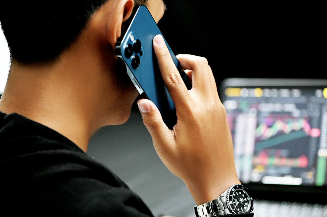 Person talking on a blue smartphone while a laptop screen shows stock charts in the background.