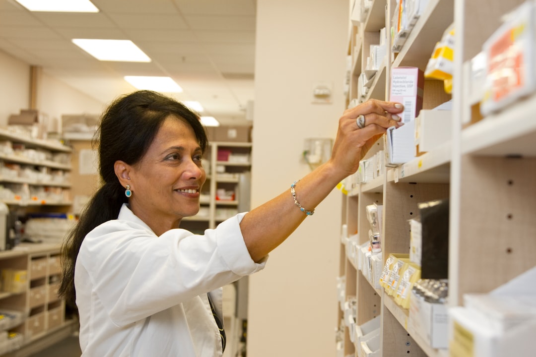 Pharmacist in a white coat reaching for a box on high shelves in a pharmacy aisle.