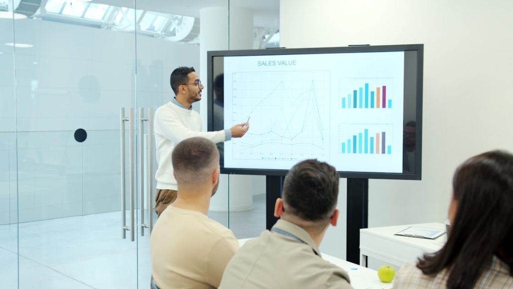 A man presenting sales data on a large screen to a small group in a bright conference room