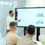 A man presenting sales data on a large screen to a small group in a bright conference room