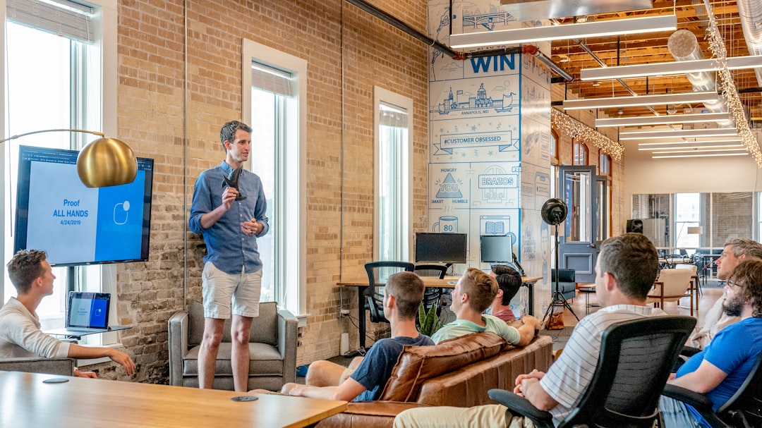 Presenter speaks to a small audience in a modern coworking space with brick walls and large windows, a screen behind him shows 'Proof ALL HANDS'.
