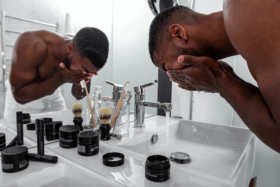 Man washing his face at a white bathroom sink with his reflection visible in the mirror, skincare jars and brushes on the counter nearby.