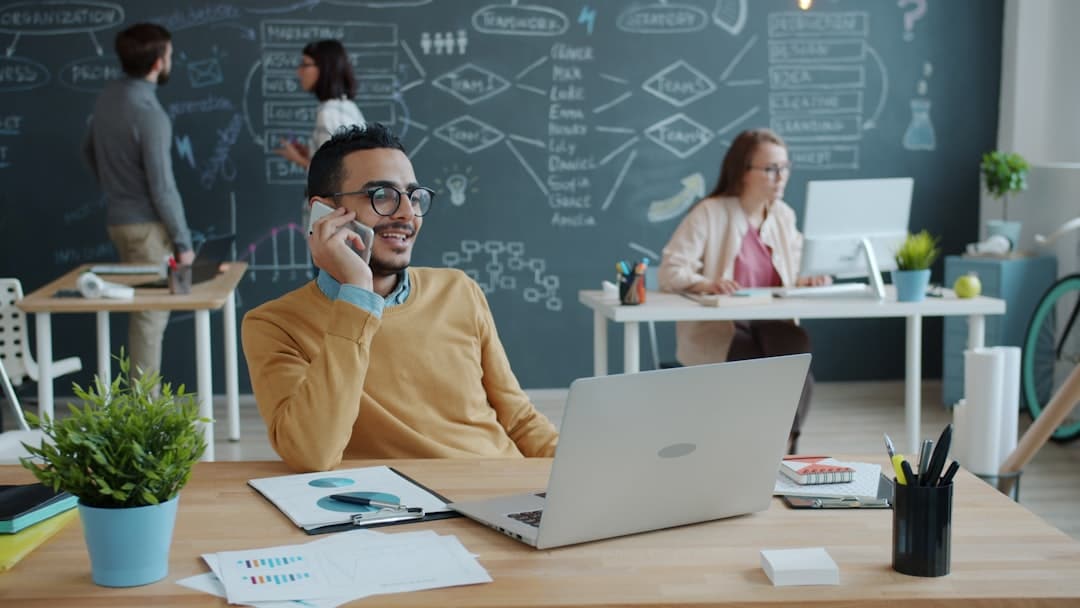 Man in a mustard sweater on a call at a desk with a laptop, papers, and a plant in a bright open-office setting.