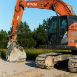 Orange Hitachi excavator with a large bucket resting on dirt at a construction site, clear blue sky behind and trees in the background.
