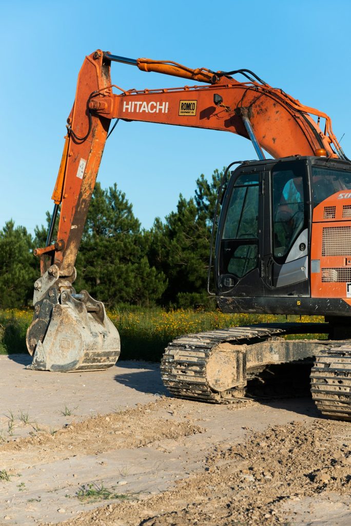 Orange Hitachi excavator with a large bucket resting on dirt at a construction site, clear blue sky behind and trees in the background.