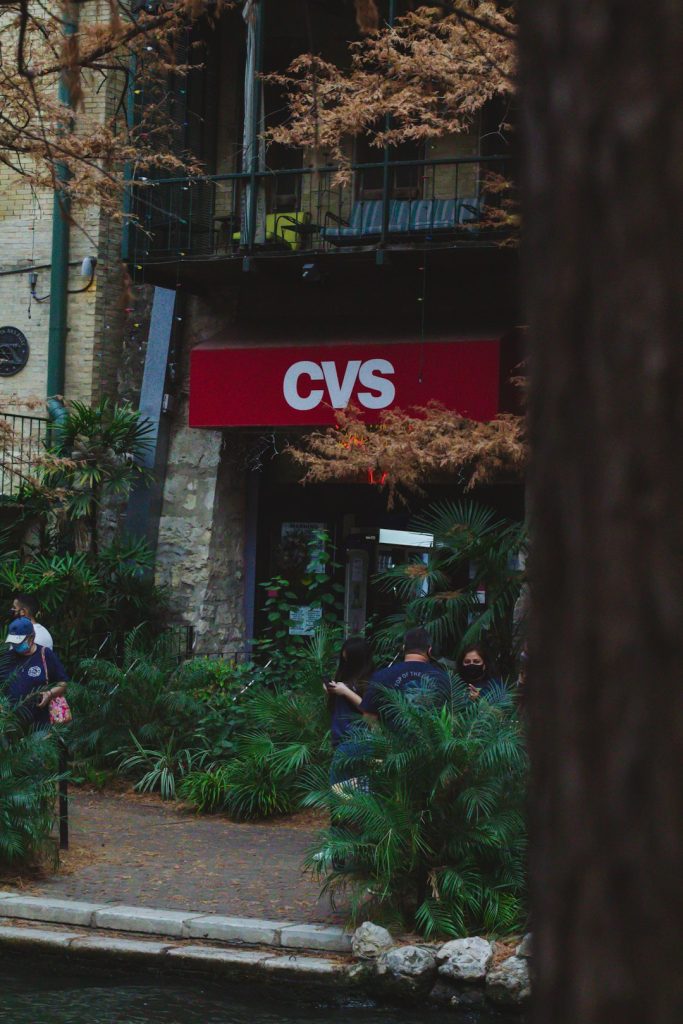CVS storefront with a red sign, people standing outside near lush plants and trees in an urban setting.