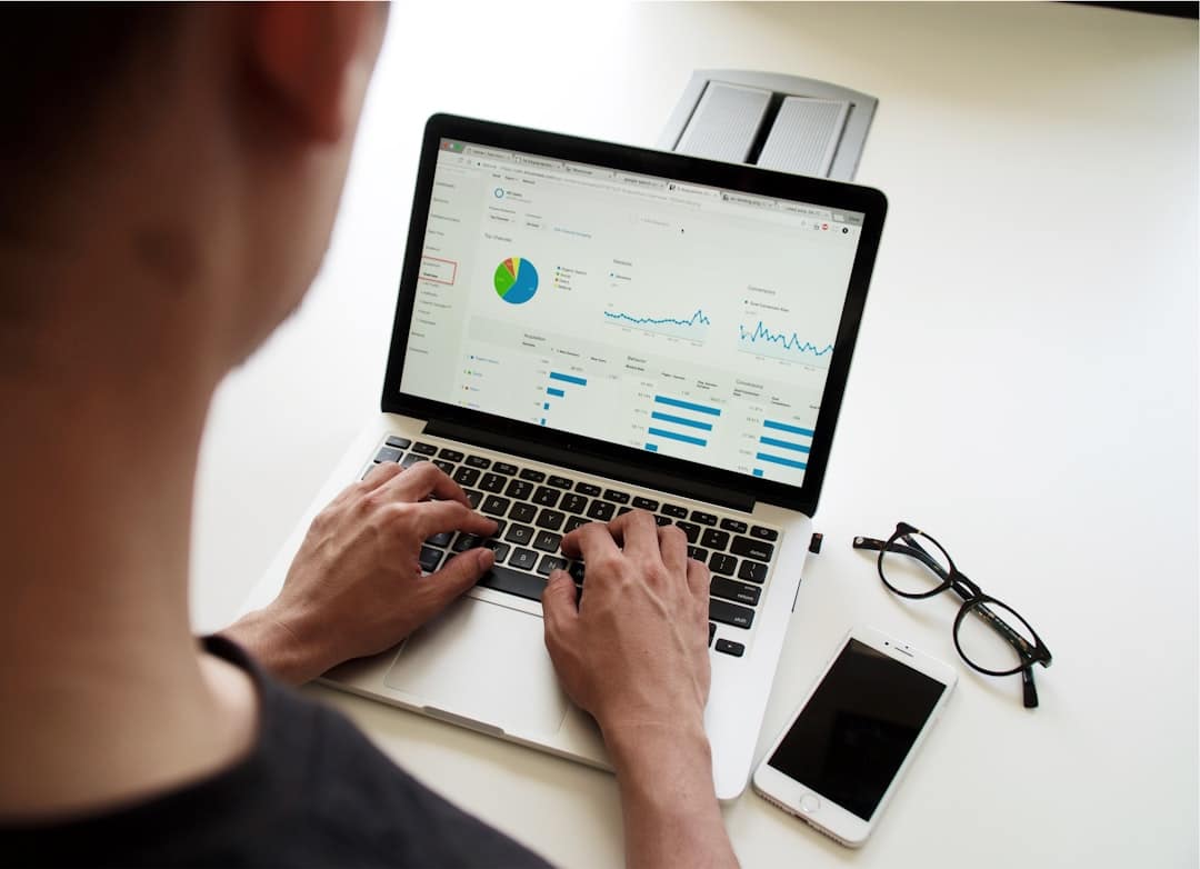 Person typing on a laptop displaying analytics dashboards with charts and graphs on a bright desk, smartphone and glasses nearby