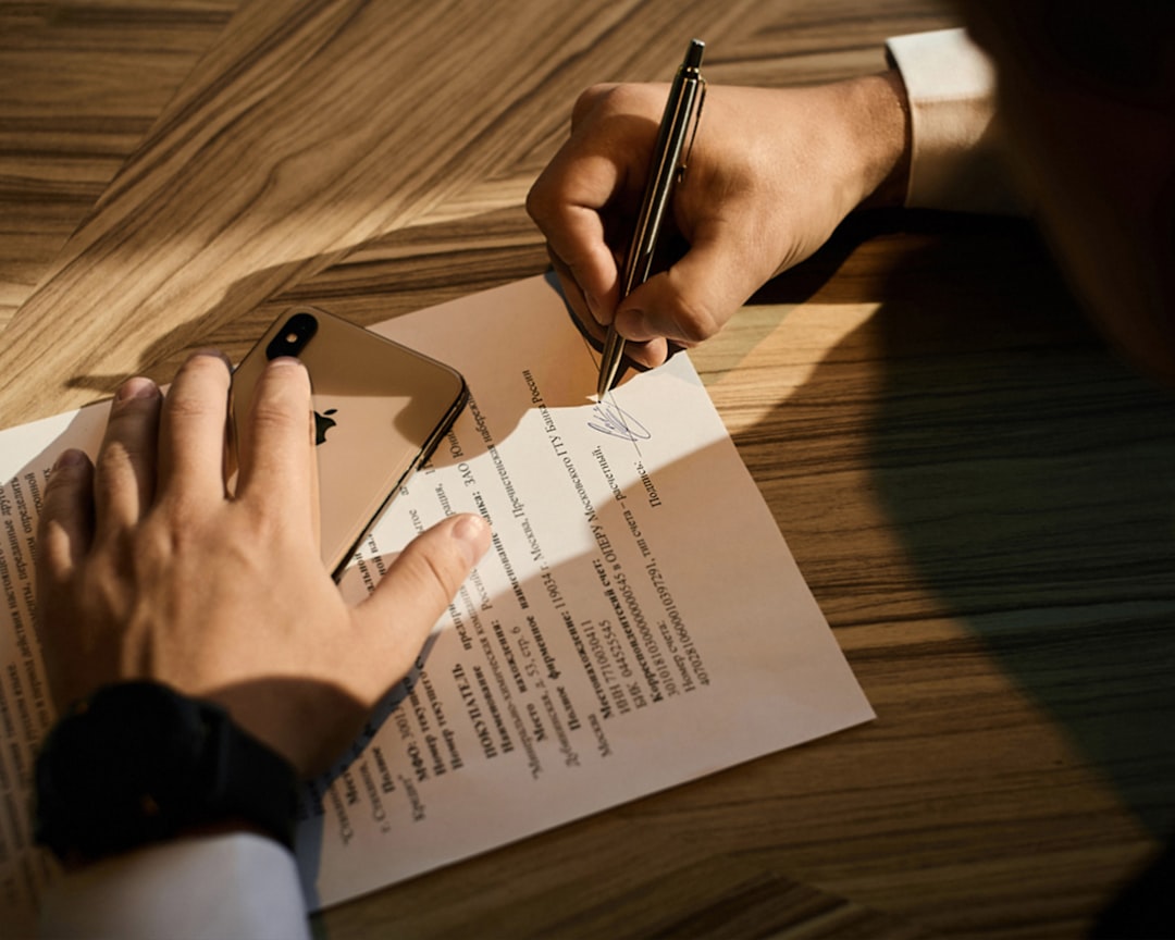 Person signs a document with a pen while resting a smartphone on the paper on a wooden table