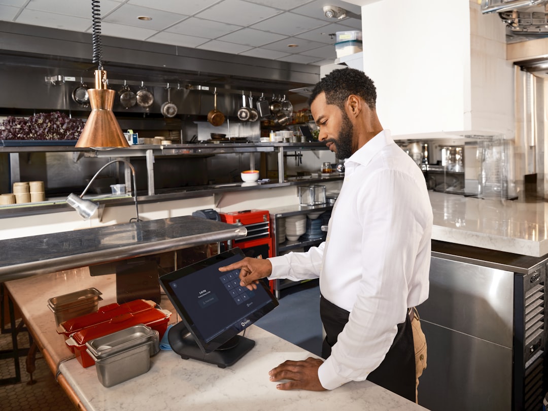 Male server in a white shirt uses a touchscreen point-of-sale terminal at a marble counter in a busy kitchen area.