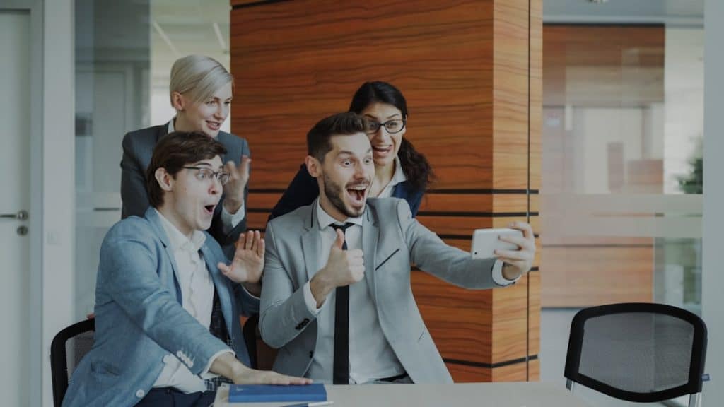 Four coworkers gather around a man in a gray suit taking a selfie, all smiling and cheering in a bright modern office.