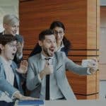 Four coworkers gather around a man in a gray suit taking a selfie, all smiling and cheering in a bright modern office.