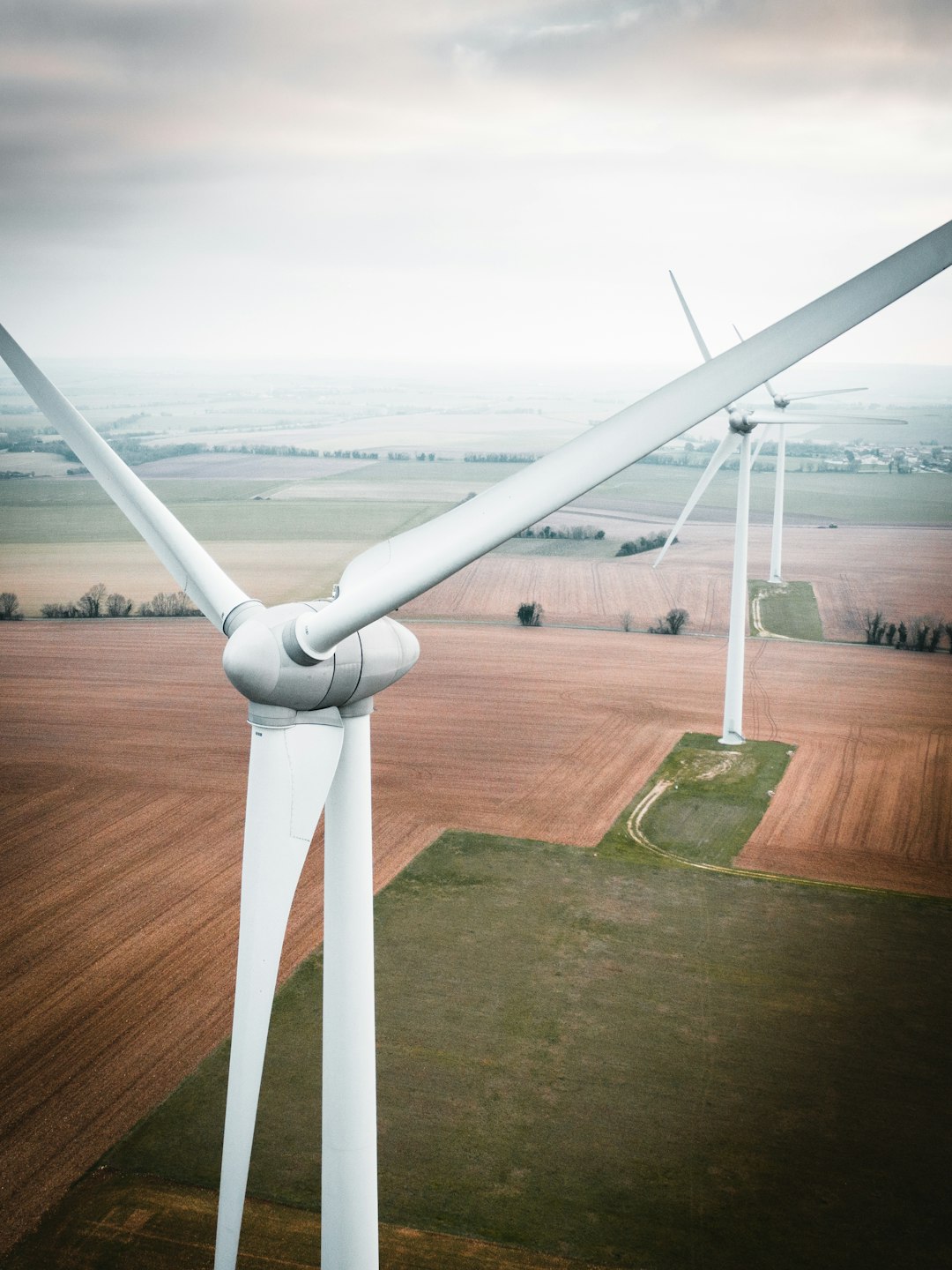 Wind turbines towering over brown and green farmland under a cloudy, pale sky, generating renewable energy