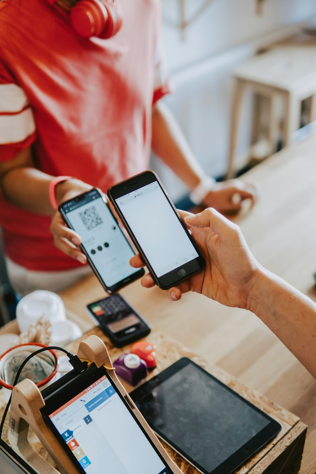 Two people compare phones at a café, one screen showing a QR code for a payment or pass, with a tablet and devices on the table nearby.