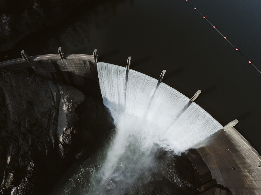 Aerial view of a curved dam spilling water over its edge into a rocky gorge below.