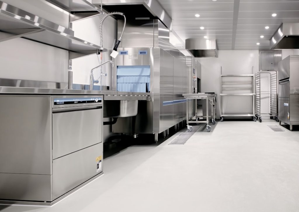 Commercial stainless steel kitchen with an industrial dishwasher, sink, and prep tables in a bright, clean space.