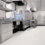 Commercial stainless steel kitchen with an industrial dishwasher, sink, and prep tables in a bright, clean space.