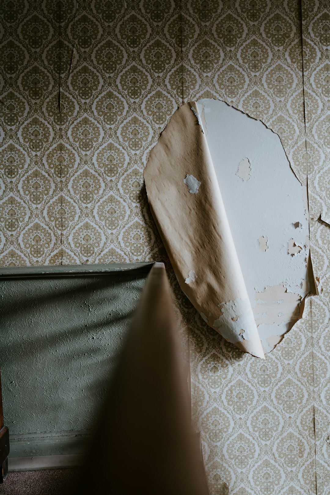 Corner of a room with ornate patterned wallpaper peeling away, revealing the bare wall and a curved, peeled panel nearby.