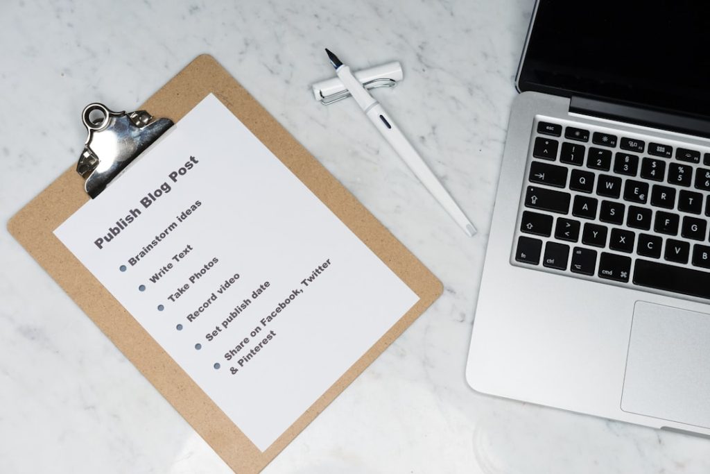 Clipboard holds a blog post checklist on a marble desk, with a white pen and a nearby laptop.