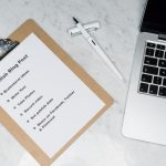 Clipboard holds a blog post checklist on a marble desk, with a white pen and a nearby laptop.