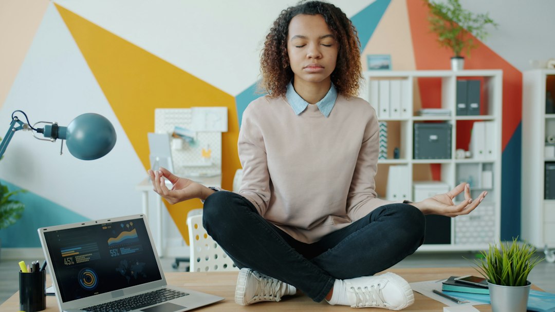 Person sits cross‑legged on the floor meditating with eyes closed in a bright, modern office setting.