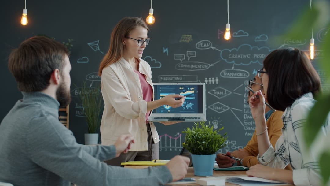 Woman presenter in a beige jacket shows a laptop with charts to three colleagues in a modern meeting room with a chalkboard background.