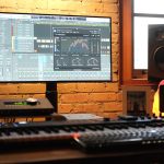 Music production desk with a large monitor showing a digital audio workstation, plus a MIDI keyboard and audio gear on a brick-walled wall setup.