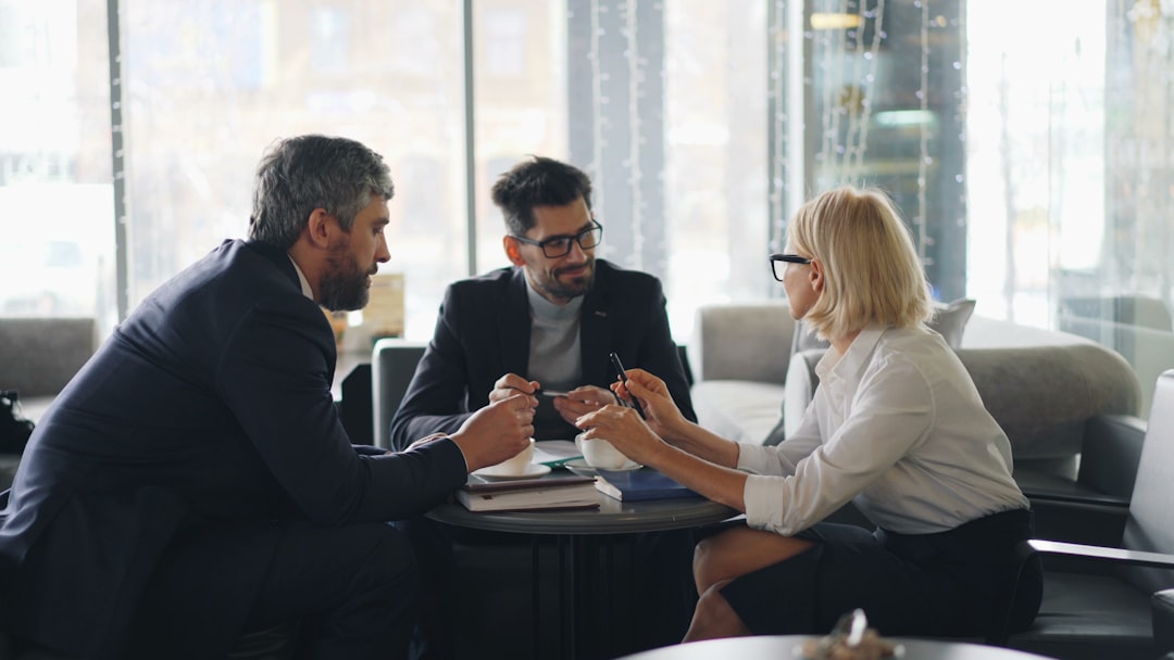 Three professionals in business attire sit around a round table in a bright, glass-walled lounge, discussing documents over coffee.