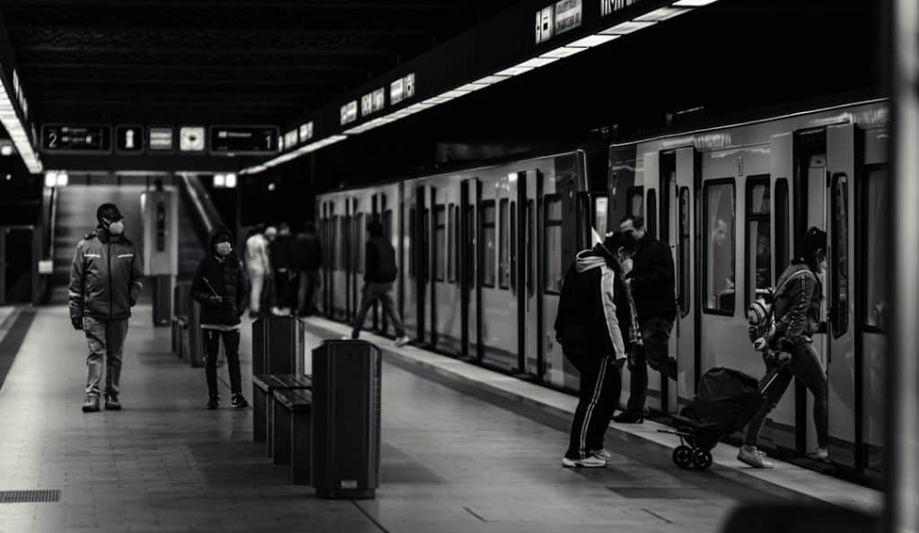 Masked commuters wait on a dimly lit subway platform as doors open on the train at right edge of the frame, a man in a jacket and a child with a suitcase nearby.