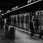 Masked commuters wait on a dimly lit subway platform as doors open on the train at right edge of the frame, a man in a jacket and a child with a suitcase nearby.