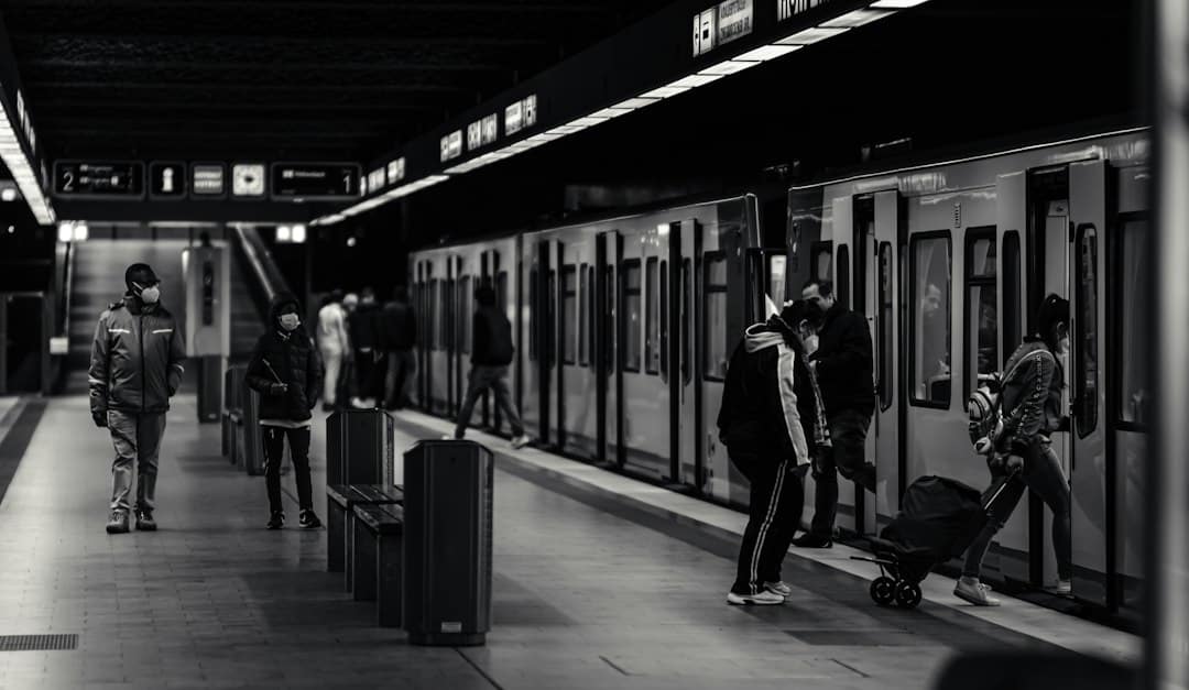 Masked commuters wait on a dimly lit subway platform as doors open on the train at right edge of the frame, a man in a jacket and a child with a suitcase nearby.