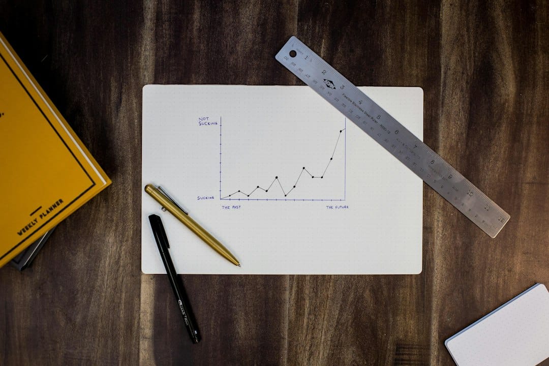 Hand-drawn line graph on a white notebook page, with a ruler across the top and pens nearby on a wooden desk.