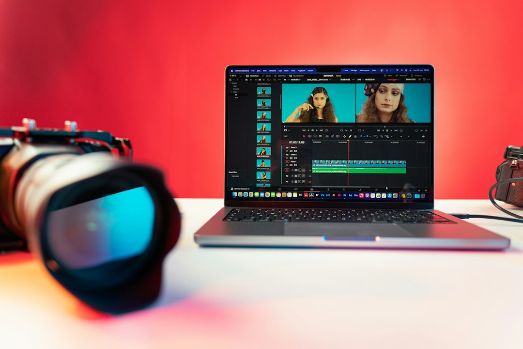 Laptop displaying video editing software with a multi-track timeline and preview panels, set against a red background with a camera lens in the foreground.