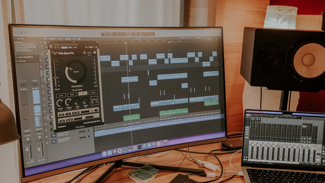 Home studio workspace with a large monitor showing a DAW timeline, a plugin window, and a laptop with a mixer app on a wooden desk beside a studio monitor speaker.
