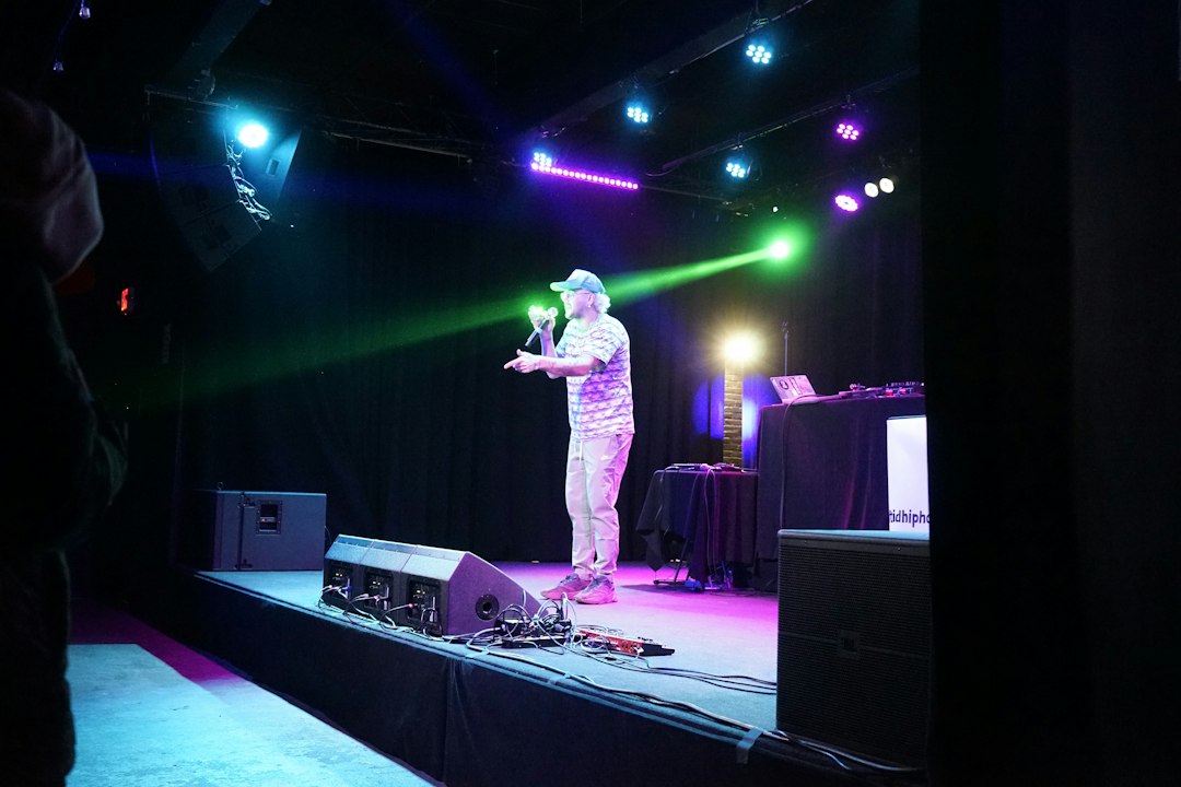 A male singer in a cap performs on a lit stage with a microphone and colorful stage lights in blue, green, and purple hues.