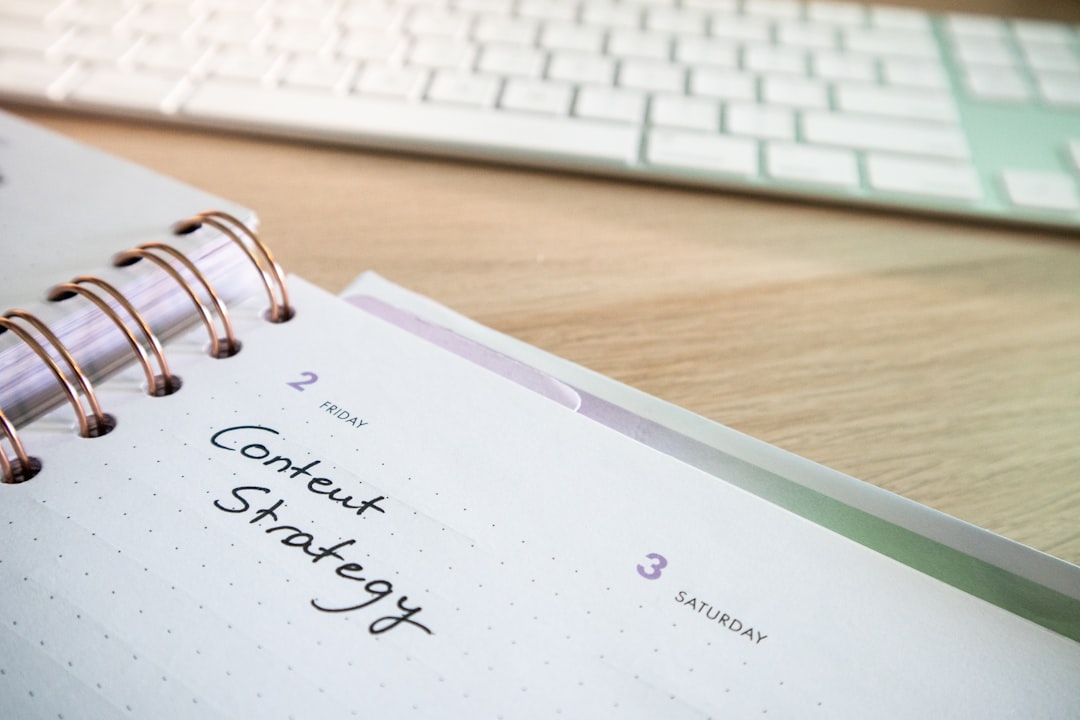 Close-up of a spiral-bound notebook on a wooden desk with 'Content Strategy' handwritten, and a keyboard blurred in the background.