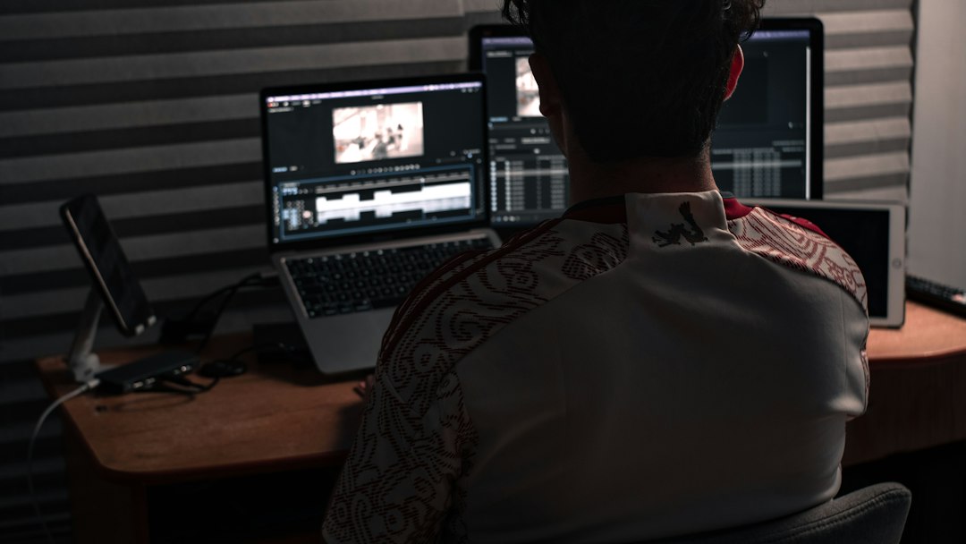 Back view of a person at a desk editing video on a laptop with external monitors in a dim studio setting.
