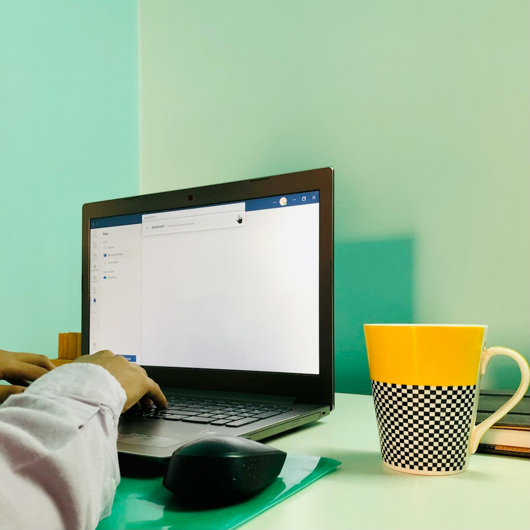Person typing on a laptop at a mint-green desk with a yellow mug nearby.