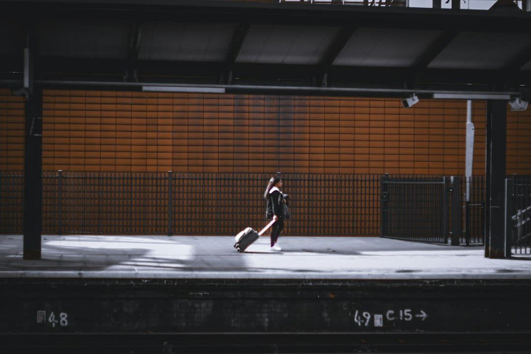 Person walking on a train platform pulling a rolling suitcase beside an orange brick wall.