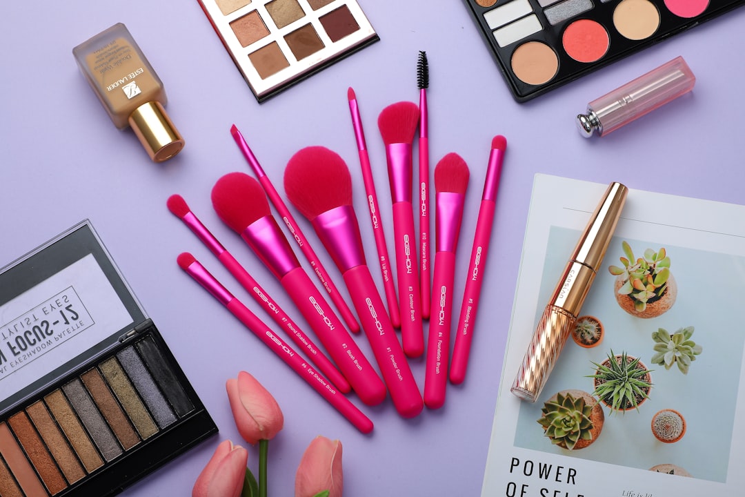 Flat lay of bright pink makeup brushes with palettes, nail polish, and lipstick on a lilac background