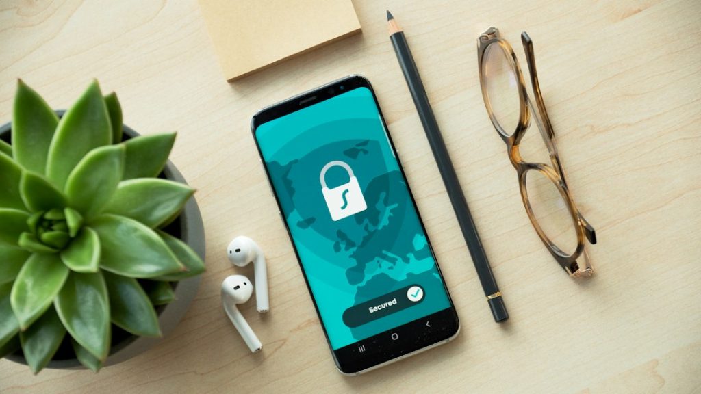 Phone screen shows a world map with a white padlock and a 'Secured' badge, illustrating mobile security on a wooden desk.