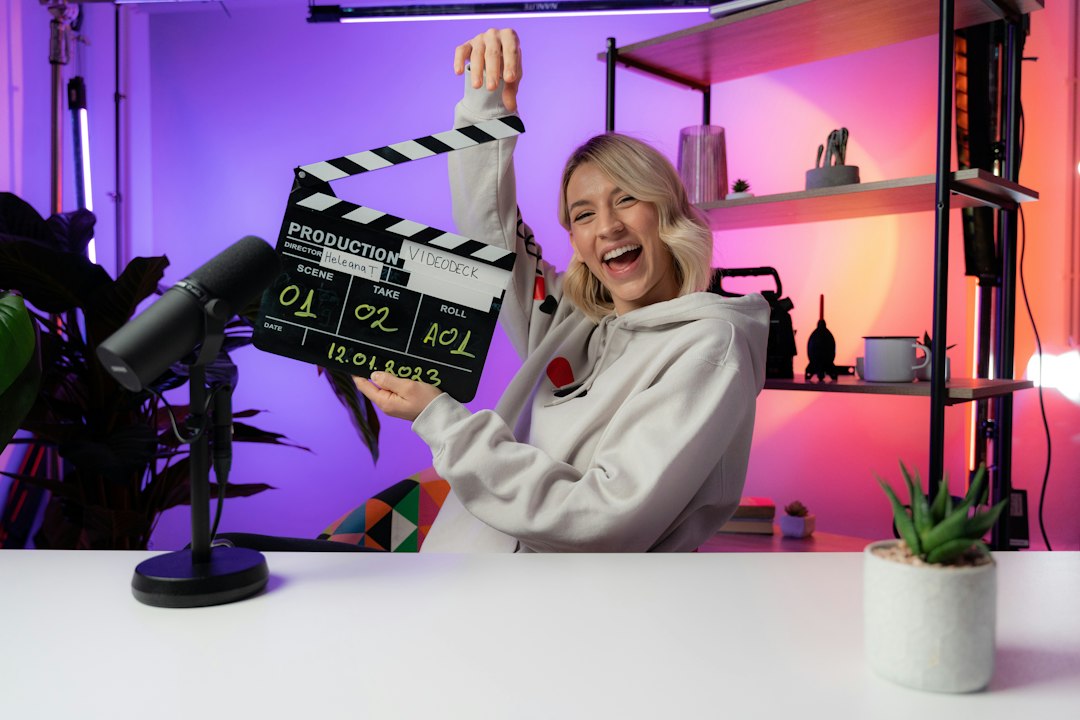 Smiling woman in a gray hoodie holds a film clapperboard in a bright podcast/film studio with purple and pink lighting, microphone in the foreground.