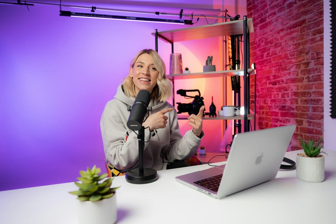 Woman with blonde hair recording a podcast at a white desk with microphone, laptop, and colorful backlighting.