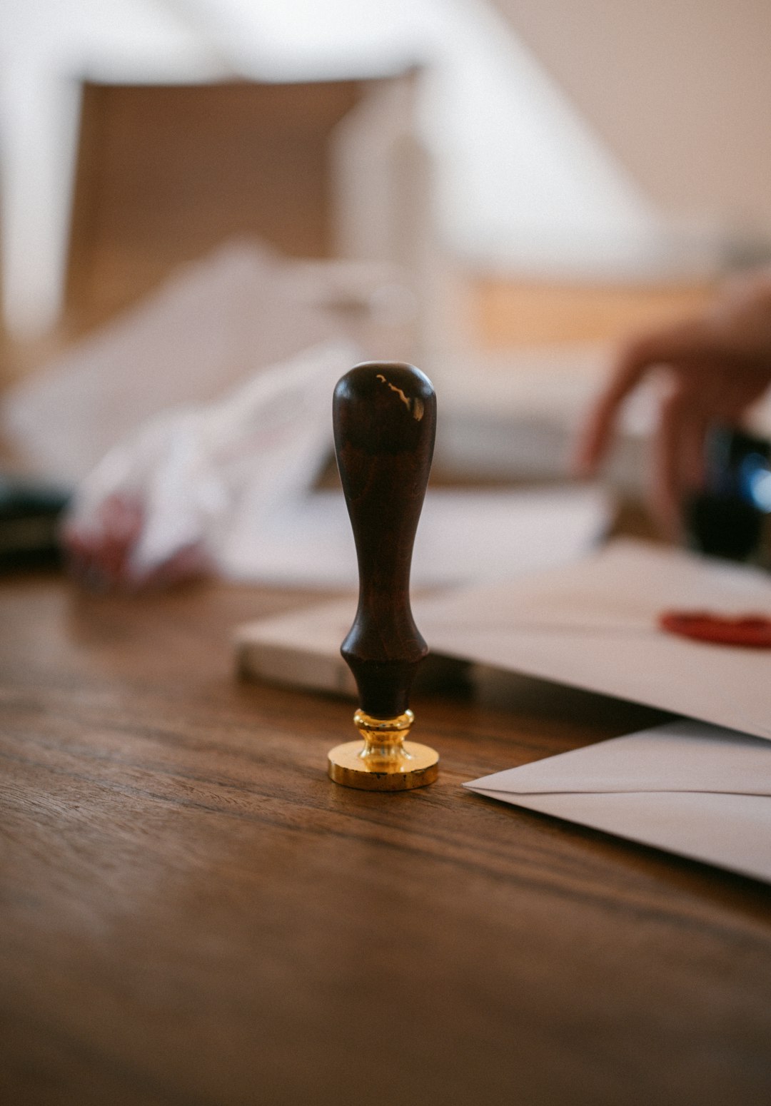 Close-up of a dark wood-handled stamp with a gold base on a wooden desk, with envelopes and papers blurred in the background.
