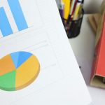 Blue bar chart and a colorful pie chart on a desk, with a red binder and pen holder nearby.