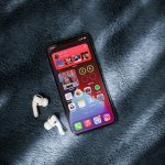 Smartphone on a blue plush blanket with white wireless earbuds beside it, screen showing colorful app icons and widgets.