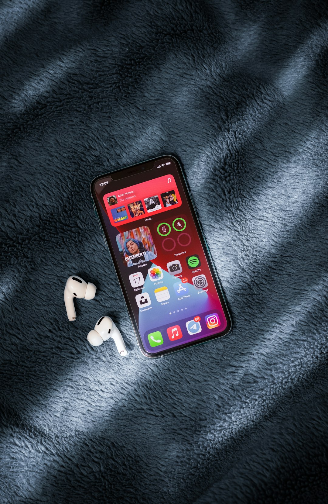 Smartphone on a blue plush blanket with white wireless earbuds beside it, screen showing colorful app icons and widgets.
