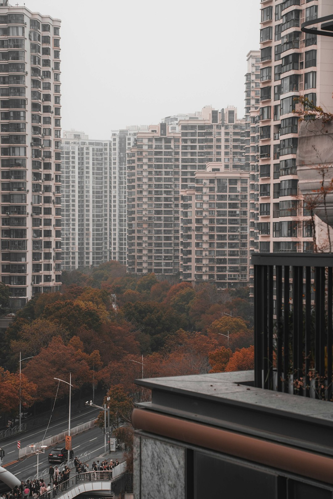 Tall, uniform apartment towers surround a central park with autumn-colored trees, viewed from a balcony over a street below on an overcast day.
