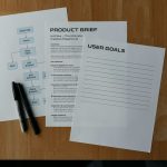 Three white sheets on a wooden desk, including a Product Brief, a User Goals page, and a flowchart with Open App/Home labels, with two markers nearby.
